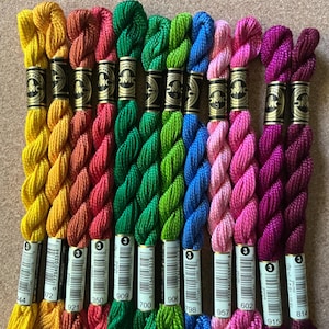 May include: A collection of ten DMC Coton Perlé size 3 embroidery thread skeins arranged in a row. The colors include yellow, orange, red, two shades of green, blue, two shades of pink, and two shades of purple. Each skein features a black and gold label.