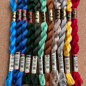 May include: A collection of ten skeins of DMC Perle cotton embroidery thread arranged vertically in a row. The colors include bright blue, dark blue, forest green, olive green, mint green, brown, light gray, dark gray, yellow, and red.
