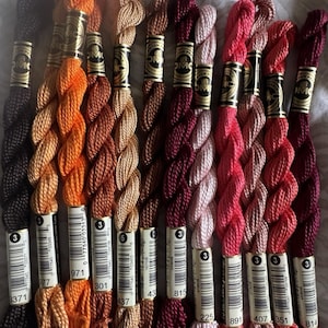 May include: A collection of eleven twisted skeins of DMC embroidery thread in various shades of black, orange, brown, pink, and deep red. Each skein features a gold paper band with the DMC logo and a white label with a barcode and color number.