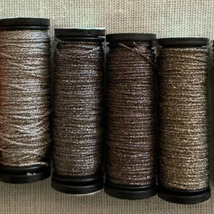 May include: Six spools of thread in various colors, including orange, silver, and brown. The thread is tightly wound on black spools, creating a textured appearance. The spools are arranged side by side.