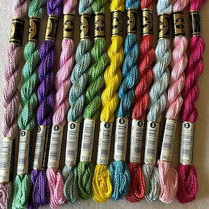 May include: A collection of DMC embroidery floss in various colors, including pink, purple, green, yellow, and blue. Each skein is labeled with a number and the DMC logo. The floss is neatly arranged, showcasing a spectrum of hues for crafting projects.