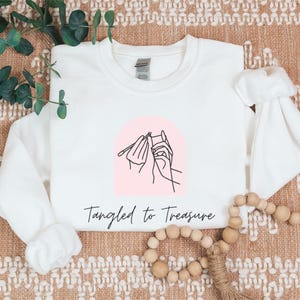 May include: White long-sleeved sweatshirt with a pink arch design featuring a line drawing of hands knitting. The text "Tangled to Treasure" is written in a script font below the design. The sweatshirt is displayed on a textured, neutral-toned surface.