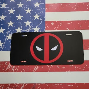 May include: Black and red license plate featuring a red Deadpool logo with white eyes. The license plate is on a red, white, and blue American flag background.