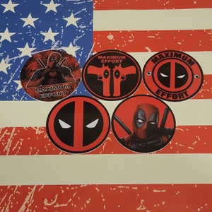 May include: Four round stickers featuring a red and black Deadpool character with the text "Maximum Effort" in black. The stickers are on a red, white, and blue American flag background.