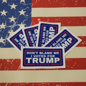May include: A stack of white stickers with blue and red text that reads "Don't Blame Me I Voted For Trump". The stickers are on top of an American flag.