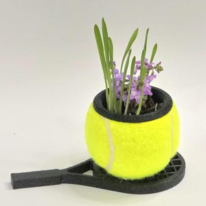 May include: A bright yellow tennis ball planter with a black rim, holding green grass and purple flowers. The planter sits on a black tennis racket-shaped base. A unique decorative item for sports enthusiasts.