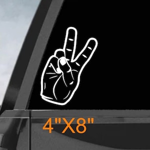 May include: White peace sign hand gesture decal sticker on a black truck tailgate. The sticker is 4 inches by 8 inches.