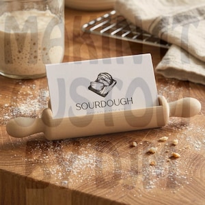 May include: A wooden rolling pin with a slot holding a business card. The card features the word "SOURDOUGH" and a bread illustration. The scene is set on a wooden surface with flour and breadcrumbs, suggesting a baking theme.