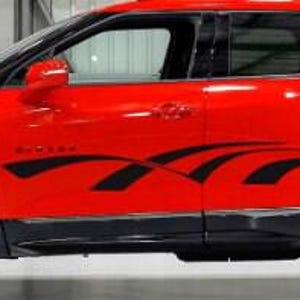 May include: A red SUV with black wheels and trim. The vehicle has a black graphic design on the side and a black roof. The car is parked in a garage or studio setting.