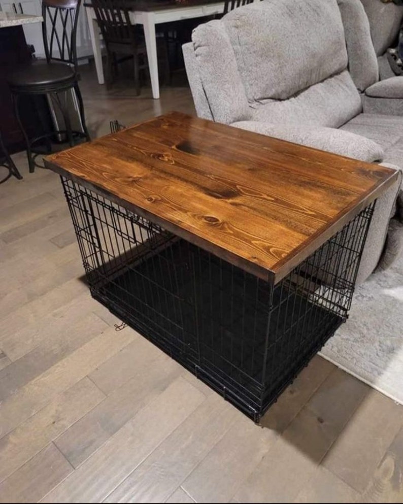 May include: A black metal dog crate with a wooden top. The crate is sitting on a wooden floor. The wooden top is stained a dark brown color.