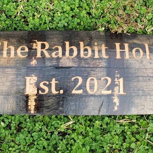 May include: A wooden sign made from a reclaimed barrel stave with the text "The Rabbit Hole Est. 2021" burned into the wood. The sign has two metal bands with gold-colored rivets.