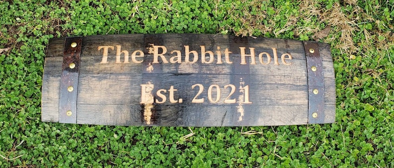 May include: A wooden sign made from a reclaimed barrel stave with the text "The Rabbit Hole Est. 2021" burned into the wood. The sign has two metal bands with gold-colored rivets.