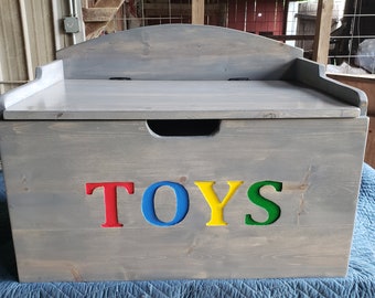 May include: A gray wooden toy box with a hinged lid and a cutout for a handle. The word "TOYS" is spelled out in colorful letters on the front of the box.