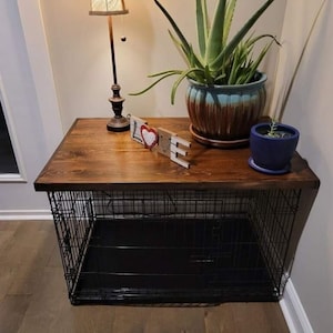 May include: A black metal dog crate with a wooden top. The crate is sitting on a wooden floor. There is a lamp, a plant, and a small wooden sign that says "LOVE" on the top of the crate.