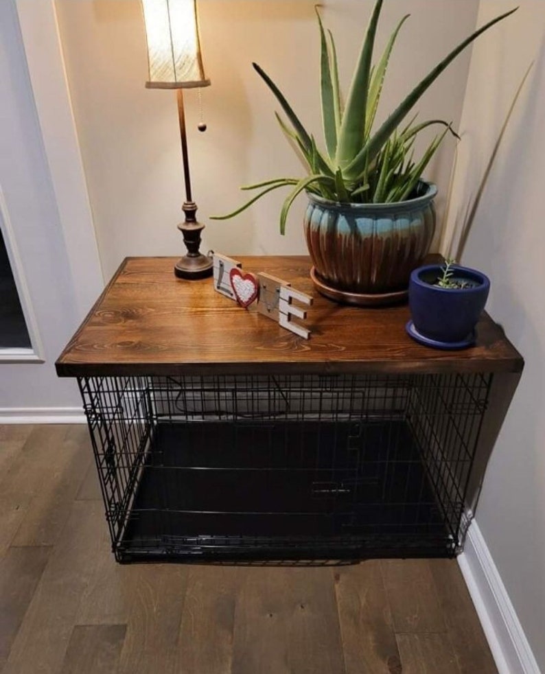 May include: A black metal dog crate with a wooden top. The crate is sitting on a wooden floor. There is a lamp, a plant, and a small wooden sign that says "LOVE" on the top of the crate.