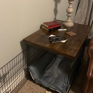 May include: A dark brown wooden table top with a black metal dog crate underneath. The table top has a few items on it, including a stack of books, a remote control, and a small decorative object. The crate has a blue dog bed inside.