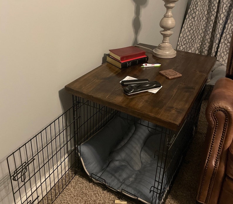 May include: A dark brown wooden table top with a black metal dog crate underneath. The table top has a few items on it, including a stack of books, a remote control, and a small decorative object. The crate has a blue dog bed inside.