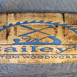 May include: A wooden sign with a dark brown stain and a blue painted logo. The logo is a saw blade with crossed tools inside. The text "Bailey's Custom Woodworking" is below the logo.