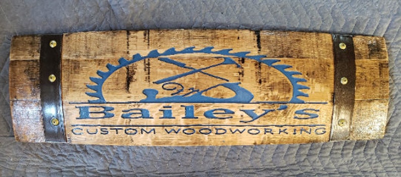May include: A wooden sign with a dark brown stain and a blue painted logo. The logo is a saw blade with crossed tools inside. The text "Bailey's Custom Woodworking" is below the logo.