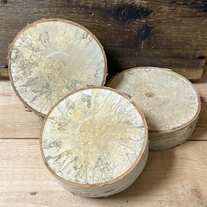 May include: Three round, natural wood coasters with a light beige and gray pattern. The coasters are made from cross-sections of a tree branch, showcasing the wood grain and bark edges. The coasters are approximately 4 inches in diameter.