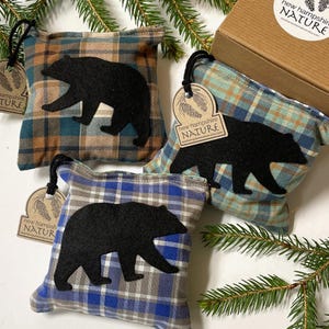 May include: Three plaid fabric sachets with black bear silhouettes. The sachets are in shades of blue, brown, and green. Each sachet has a tag that reads "new hampshire NATURE". A cardboard box with a label is also visible.