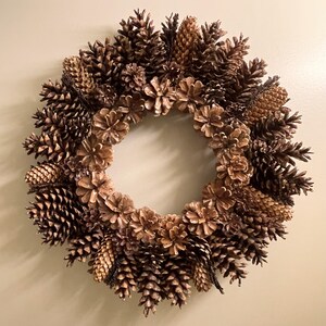 May include: A brown pine cone wreath with a variety of sizes and shapes of pine cones. The wreath is made of natural materials and has a rustic look.
