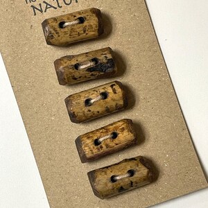 May include: Five rectangular wooden buttons with two holes each, strung with white thread, are displayed on a brown card. The buttons have a natural, rustic appearance with variations in wood grain and color.