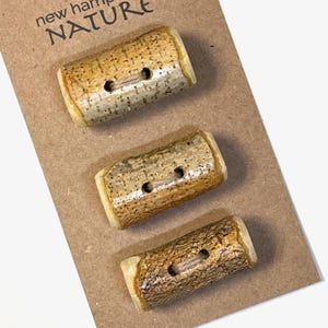 May include: Three rustic, wooden buttons with a bark-like texture and two holes each. The buttons are rectangular with rounded edges and are attached to a brown card. The card has the words "new hampshire NATURE" printed at the top.