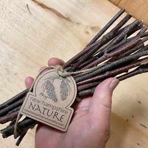 May include: A bundle of brown and reddish-brown sticks tied together with twine. A brown paper tag with the text "new hampshire NATURE newhampshirenature.etsy.com" is attached to the bundle.
