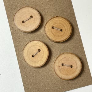 May include: Four circular buttons crafted from natural wood slices, each featuring visible grain patterns and two central holes connected by a thin piece of light-colored string. The buttons are arranged in a cluster on a flat, textured brown surface.