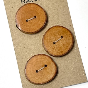 May include: Three round wooden buttons with a natural wood grain finish. Each button has two holes and is attached with a beige thread. The buttons are displayed on a brown card.