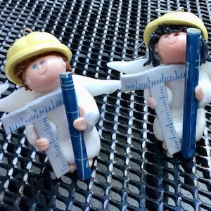 May include: Five white angel ornaments with yellow hard hats and blue tools. The tools include a ruler, a protractor, and a pencil. The angels are holding the tools in their hands.