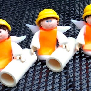 May include: Three small angel figurines wearing yellow hard hats and orange safety vests. Each angel is holding a white pipe in their hands.