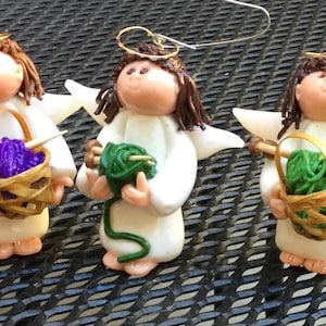 May include: Five white angel figurines with gold halos, each holding a basket of yarn in different colors. The angels are wearing white dresses and have blonde or brown hair.