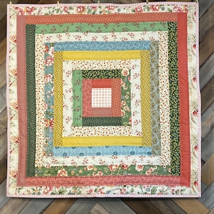 May include: Square quilt with a log cabin design. The quilt features a variety of floral and patterned fabrics in shades of green, pink, yellow, and blue. The quilt is approximately 30 inches square.