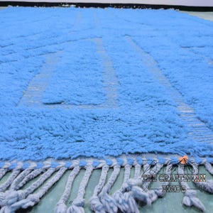 May include: A light blue rug with a geometric pattern and fringe. The rug is made of a soft, fluffy material.