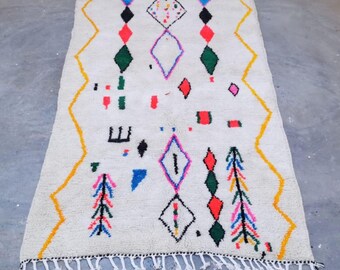 May include: A rectangular, off-white rug with a geometric pattern. The design includes colorful diamonds, triangles, and zig-zag lines in red, green, blue, and yellow. The rug has a fringe on the short ends.