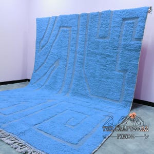 May include: A light blue rug with a geometric pattern in a lighter shade of blue. The rug has a fringe border.