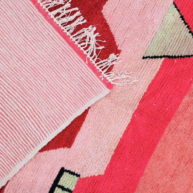 Puede incluir: Primer plano de una colorida alfombra con un dise&ntilde;o geom&eacute;trico. La alfombra presenta tonos de rojo, rosa y verde. La alfombra tiene un borde con flecos y una parte inferior a rayas. La alfombra est&aacute; hecha de un material suave y texturizado.