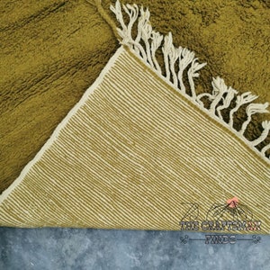 May include: Close-up of a green and white woven rug with fringe. The rug is folded in half, showing the underside of the rug.