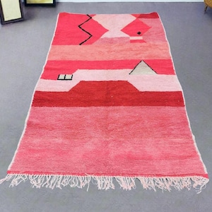 Puede incluir: Una alfombra rectangular tejida a mano en tonos rosas y rojos. La alfombra presenta formas geom&eacute;tricas y rayas horizontales, con flecos en el borde inferior. El dise&ntilde;o incluye un zigzag y un tri&aacute;ngulo negros. Esta alfombra es adecuada para la decoraci&oacute;n del hogar.