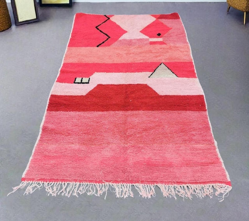 Puede incluir: Una alfombra rectangular tejida a mano en tonos rosas y rojos. La alfombra presenta formas geom&eacute;tricas y rayas horizontales, con flecos en el borde inferior. El dise&ntilde;o incluye un zigzag y un tri&aacute;ngulo negros. Esta alfombra es adecuada para la decoraci&oacute;n del hogar.
