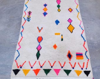 May include: A rectangular, off-white rug with a geometric design. The rug features colorful shapes including diamonds, triangles, and zigzags in shades of pink, orange, yellow, blue, and green. The rug has a fringe on the short ends.