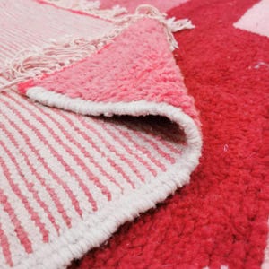 Puede incluir: Primer plano de una alfombra rosa y roja con un dise&ntilde;o geom&eacute;trico. La alfombra tiene una superficie texturizada y afelpada con un borde blanco. El dise&ntilde;o de la alfombra incluye rayas y bloques de color s&oacute;lidos.