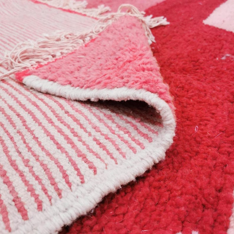 Puede incluir: Primer plano de una alfombra rosa y roja con un dise&ntilde;o geom&eacute;trico. La alfombra tiene una superficie texturizada y afelpada con un borde blanco. El dise&ntilde;o de la alfombra incluye rayas y bloques de color s&oacute;lidos.