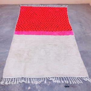 Op de afbeelding: Een rechthoekig tapijt met een colorblock-ontwerp. Het bovenste gedeelte is rood met kleine zwarte stippen, gevolgd door een roze streep en een groot gebroken wit gedeelte. Het tapijt heeft witte franjes langs de onderkant.