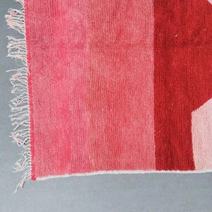 Puede incluir: Primer plano de una secci&oacute;n de una alfombra con un dise&ntilde;o geom&eacute;trico. La alfombra presenta una paleta de colores rosa y rojo. El dise&ntilde;o incluye una franja vertical y una secci&oacute;n con una forma geom&eacute;trica. La alfombra tiene flecos en un lado.