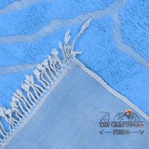 May include: Close-up of a light blue rug with a fringe border. The rug has a subtle striped pattern and a soft, fluffy texture.