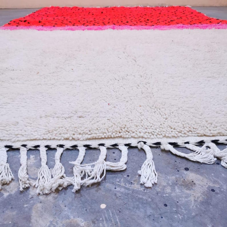 Op de afbeelding: Een rechthoekig vloerkleed met een wit middenveld, een roze en rode bovenrand en witte kwasten langs de onderrand. Het vloerkleed heeft een getextureerd, zacht oppervlak en een zwart-witte rand.