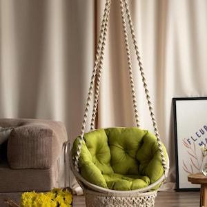 Könnte beinhalten: Ein hängender Makramee-Stuhl mit einem runden, olivgrünen Kissen. Der Stuhl ist an einem beigefarbenen Seil aufgehängt und hat unten eine dekorative Fransen. Ein Korb mit gelben Blumen steht in der Nähe.